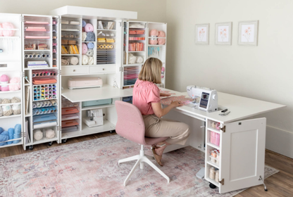 Person working at a sewing desk in a craft room with organized shelves and supplies. [Standard]
