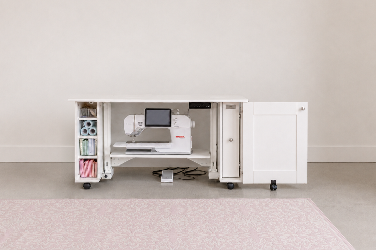 A white under-desk sewing machine cabinet with open shelves and a side cabinet door, shown in a bright room with a light pink rug. A white sewing machine is placed inside the desk. [xl]