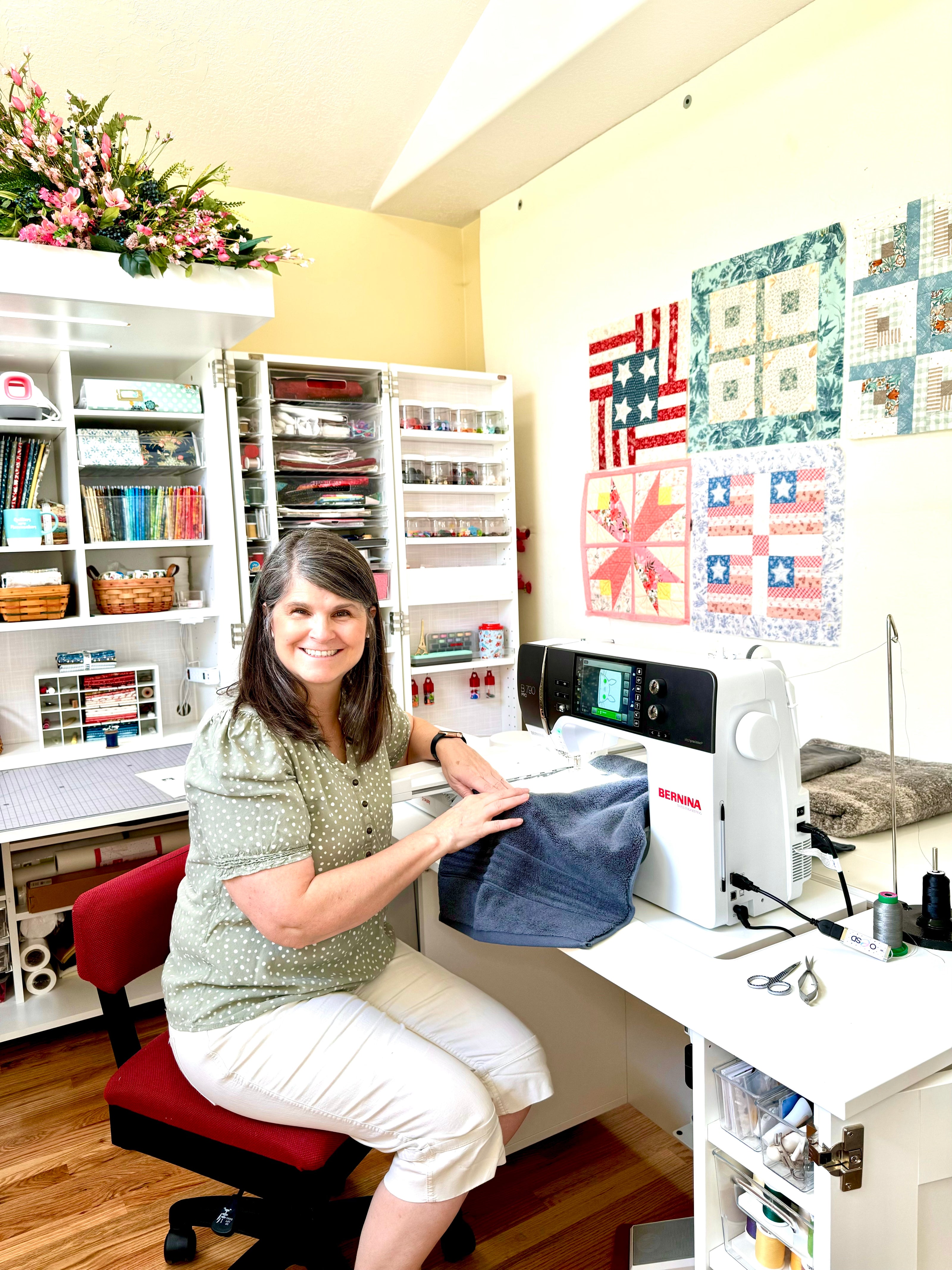 Woman sitting at a sewing machine in an organized craft room with quilts on the wall.