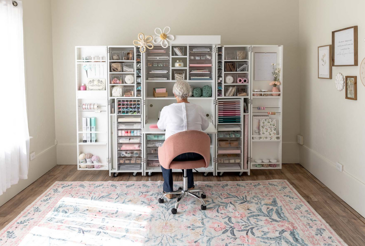 Woman sitting at a crafting station with a white storage cabinet holding well-organized craft suppli